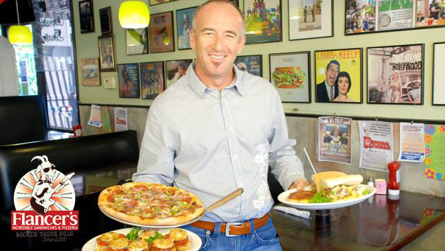 Jeff Flancer holding a tray of pizza in one hand, and a sandwich in the other. Flancer's Incredible Sandwiches & Pizzeria.